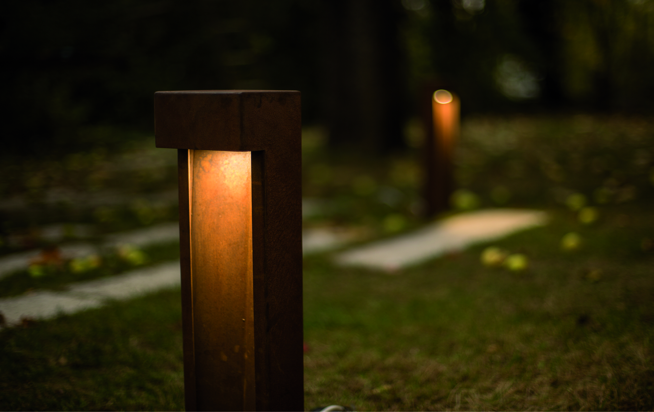 Das Bild zeigt eine Nahaufnahme einer warm beleuchteten Gartenlampe bei Dämmerung. Im Hintergrund sind weitere Lampen und ein Weg verschwommen sichtbar, umgeben von Gras und herbstlichem Laub.