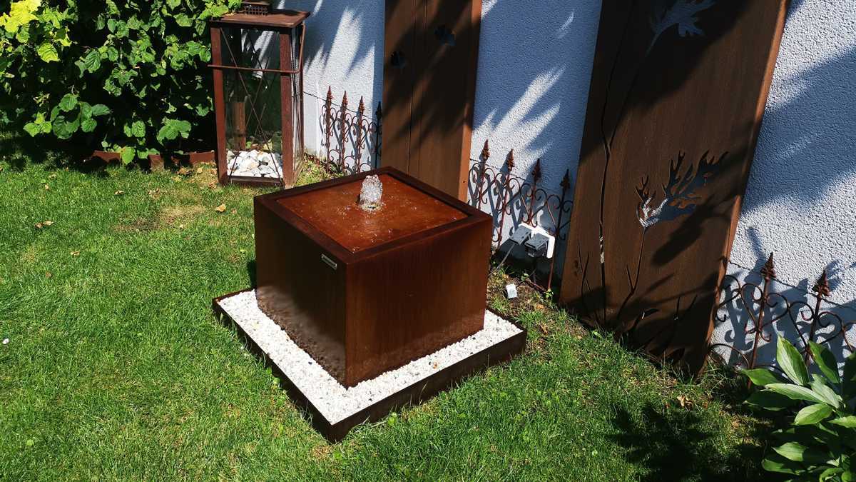 Cortenstahl Brunnen AQUA CUBUS mit Rand, in Kiesbett aufgestellt
